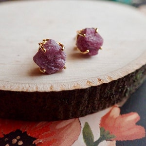 May include: A pair of stud earrings featuring raw, unpolished pinkish-purple gemstones set in gold-toned metal prongs. The stones have a jagged, natural texture and are secured by four metal claws, showcasing a rough, organic appearance.