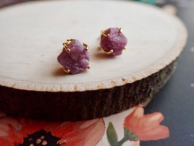 May include: A pair of stud earrings featuring raw, unpolished pinkish-purple gemstones set in gold-toned metal prongs. The stones have a jagged, natural texture and are secured by four metal claws, showcasing a rough, organic appearance.