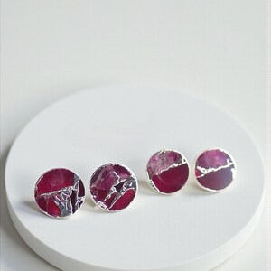 May include: Four pairs of round stud earrings with a silver border. The earrings feature a deep magenta and silver marbled design. They are displayed on a white, circular surface.