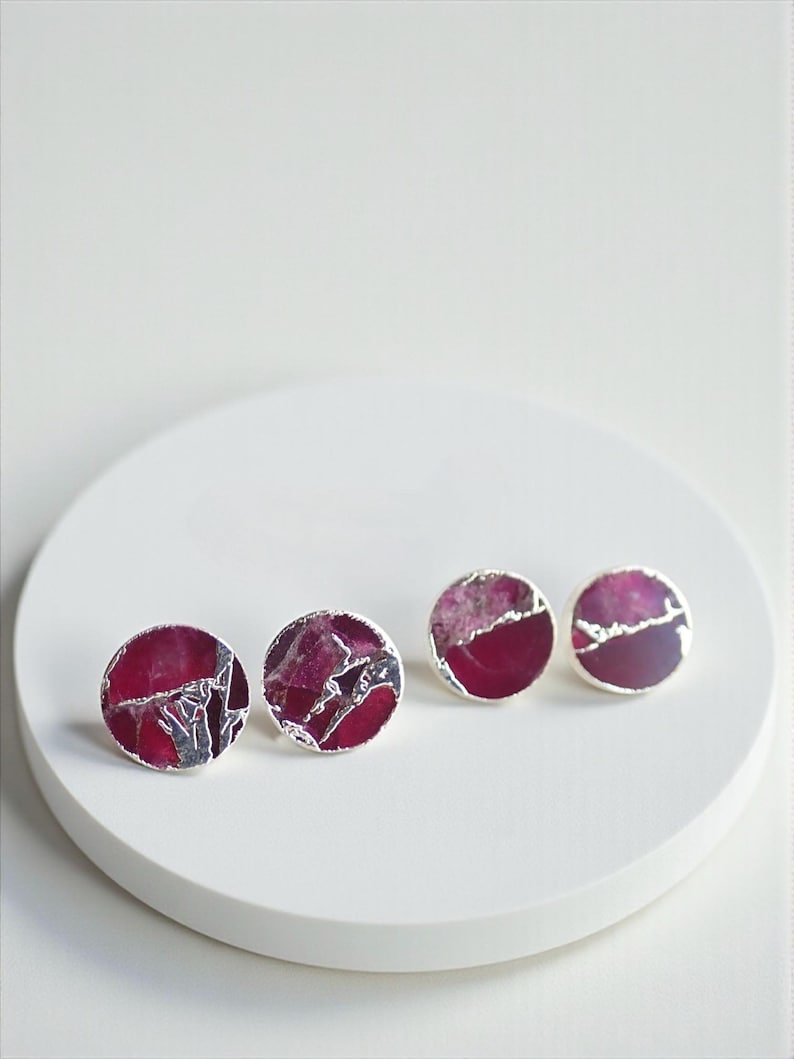 May include: Four pairs of round stud earrings with a silver border. The earrings feature a deep magenta and silver marbled design. They are displayed on a white, circular surface.