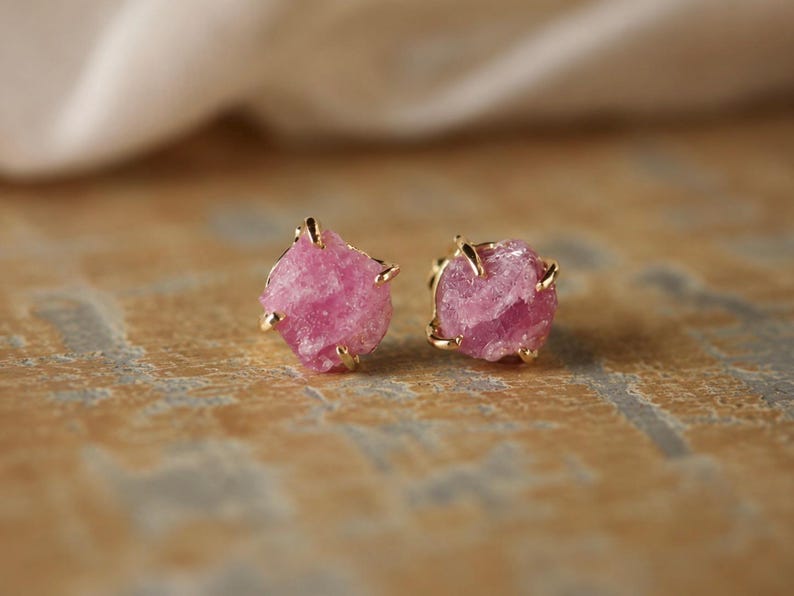 May include: A pair of stud earrings featuring raw, vibrant pink gemstones set in gold-toned metal prongs. Each earring showcases a jagged, natural stone texture with a bright magenta hue, securely held by four delicate metallic claws.