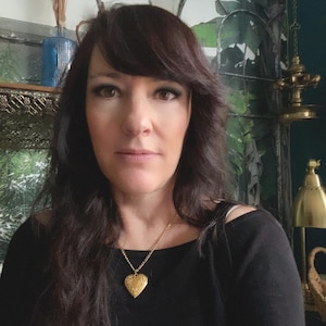 Pode incluir: Uma mulher com cabelo comprido e escuro vestindo uma camisa preta e um colar com um pingente em forma de cora&ccedil;&atilde;o de ouro.