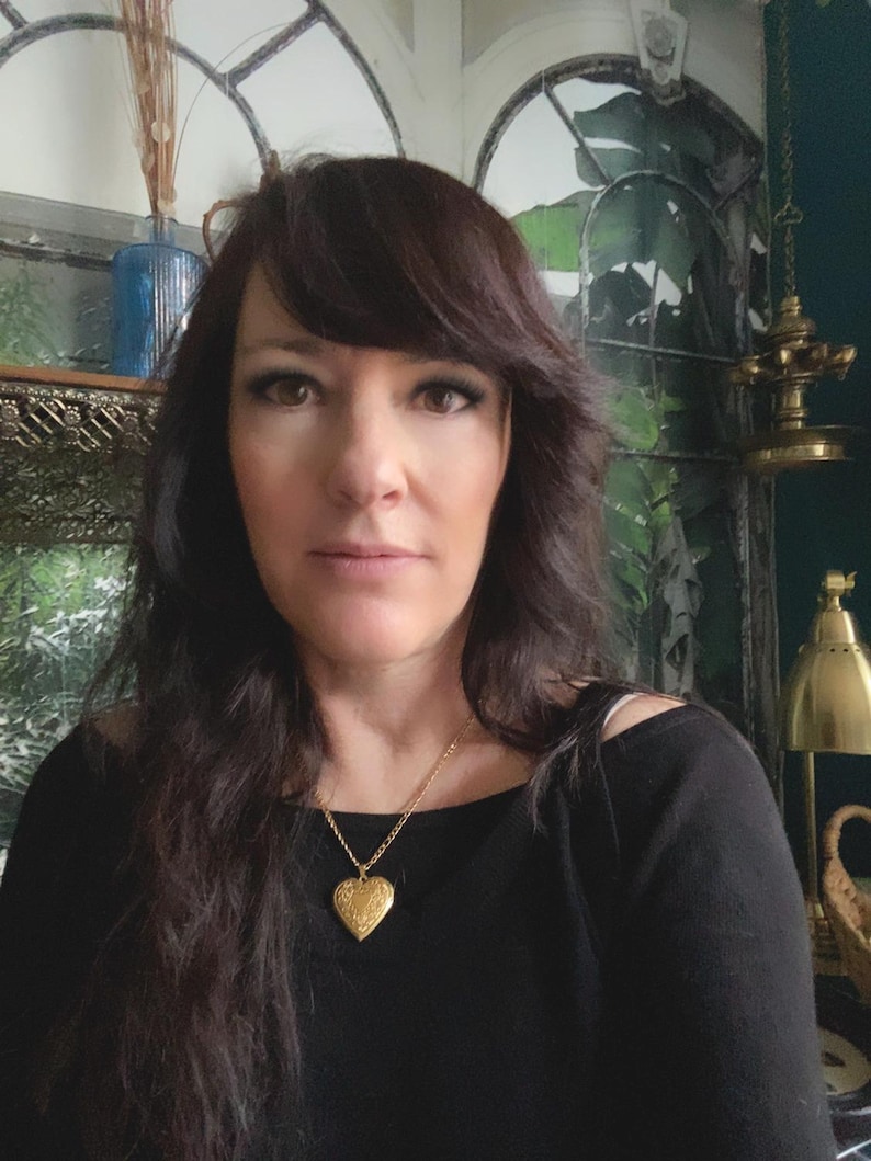 Pode incluir: Uma mulher com cabelo comprido e escuro vestindo uma camisa preta e um colar com um pingente em forma de cora&ccedil;&atilde;o de ouro.