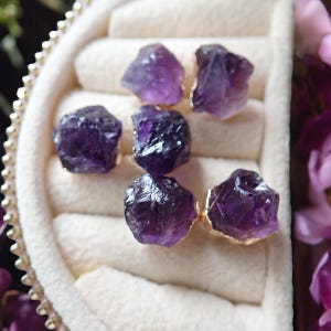 May include: Six raw amethyst gemstone earrings with gold-colored settings. The earrings are a deep purple color and are displayed in a white jewelry box. The gemstones have a rough, natural texture.