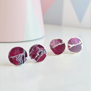 May include: Four pairs of round stud earrings with a silver border. The earrings have a deep red, marbled stone design. The background includes a pink cylinder and a geometric pattern in pink, blue, and white.