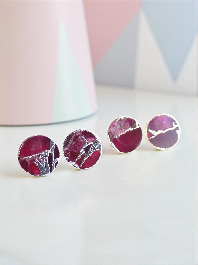 May include: Four pairs of round stud earrings with a silver border. The earrings have a deep red, marbled stone design. The background includes a pink cylinder and a geometric pattern in pink, blue, and white.