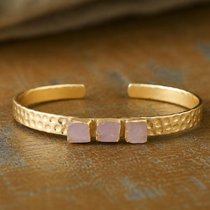 May include: A gold-toned cuff bracelet featuring a hammered texture along the band and three square-cut, pale pink stones set horizontally in the center. The bracelet has an open back design, allowing for adjustment to fit the wrist securely.