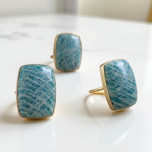 May include: Three rectangular earrings with a gold-colored frame. The earrings have a light blue stone with white veining. The earrings are displayed on a white surface.