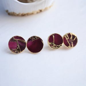 May include: Four pairs of round stud earrings with a gold border. The earrings have a deep magenta centre with gold vein-like accents. The earrings are displayed on a white surface.