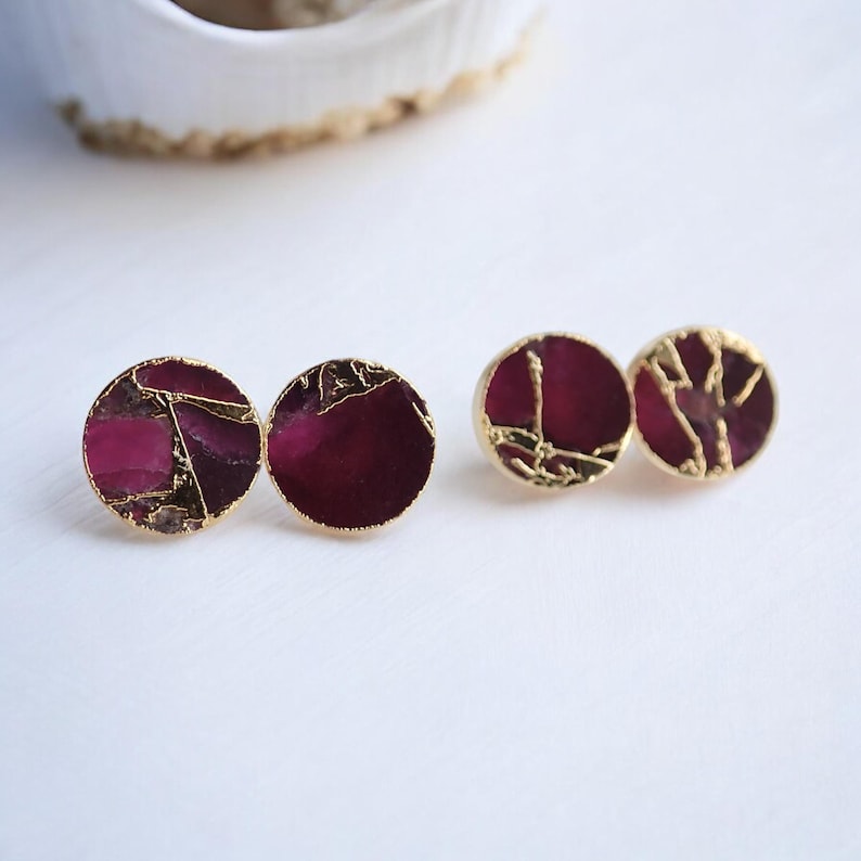 May include: Four pairs of round stud earrings with a gold border. The earrings have a deep magenta centre with gold vein-like accents. The earrings are displayed on a white surface.