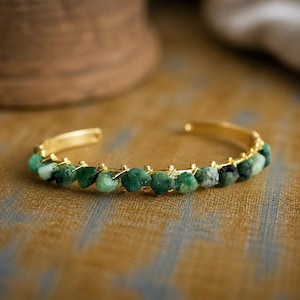 May include: A gold-toned cuff bracelet featuring a row of rough-cut, vibrant green emerald stones. The bracelet has a delicate, open design with a polished finish. The emeralds are securely set along the band, creating a natural, organic aesthetic.
