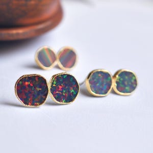 May include: Gold-plated stud earrings featuring a pair of iridescent, oval-shaped opals. The opals display a mix of red, green, and blue hues. The earrings are set against a white background, with a blurred background.