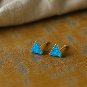 May include: A pair of gold stud earrings with triangular blue opal inlays. The earrings are set on a textured, golden-brown surface, with a neutral-colored fabric in the background. The earrings are small and delicate.