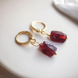 May include: A pair of gold hoop earrings with a dangling raw garnet gemstone. The garnet is a deep red color and has a natural, rough texture.