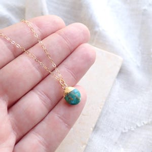 Peut inclure: Un d&eacute;licat collier &agrave; cha&icirc;ne dor&eacute;e avec un petit pendentif en pierre de couleur turquoise. La pierre a un capuchon dor&eacute;. Le collier est pr&eacute;sent&eacute; sur une surface de couleur claire.