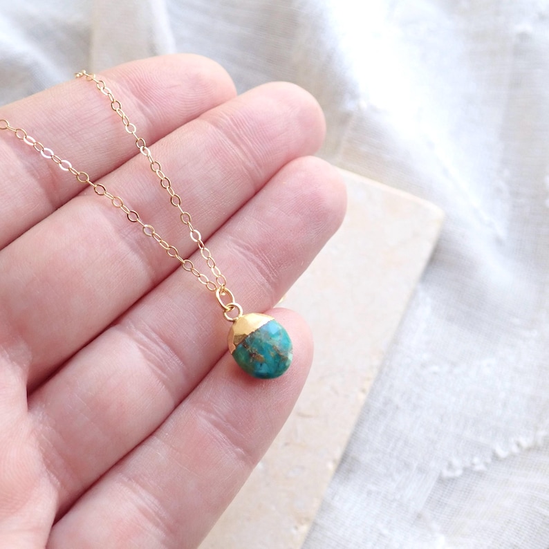 Peut inclure: Un d&eacute;licat collier &agrave; cha&icirc;ne dor&eacute;e avec un petit pendentif en pierre de couleur turquoise. La pierre a un capuchon dor&eacute;. Le collier est pr&eacute;sent&eacute; sur une surface de couleur claire.