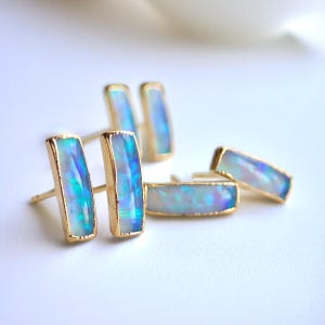 May include: Gold-plated bar stud earrings with iridescent opal stones. The opals display a range of blues and greens, set in a rectangular shape. The earrings are displayed on a white surface.