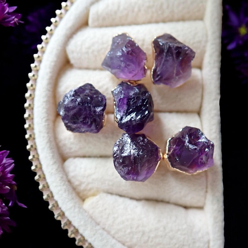 May include: Six raw amethyst gemstones with gold-colored edges are displayed in a white jewelry box. The gemstones are a deep purple color and have a rough, natural texture. The box has a soft, padded interior and a decorative gold trim.