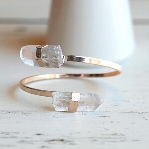 May include: A gold-toned, open cuff bracelet featuring two clear quartz crystal points. The crystals are secured with gold-colored metal bands. The bracelet has a minimalist design and is displayed on a white surface.