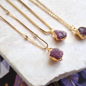 May include: Three gold chain necklaces with adjustable lengths, each featuring a different raw, purple gemstone pendant. The gemstones are irregularly shaped and have a natural, rough texture.