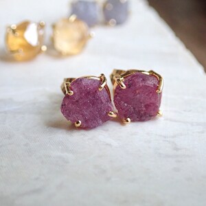 May include: A pair of stud earrings featuring raw, deep pink gemstones set in gold-toned metal prongs. In the background, additional pairs of stud earrings with yellow and light blue raw gemstones are visible, each secured with gold-toned prongs.