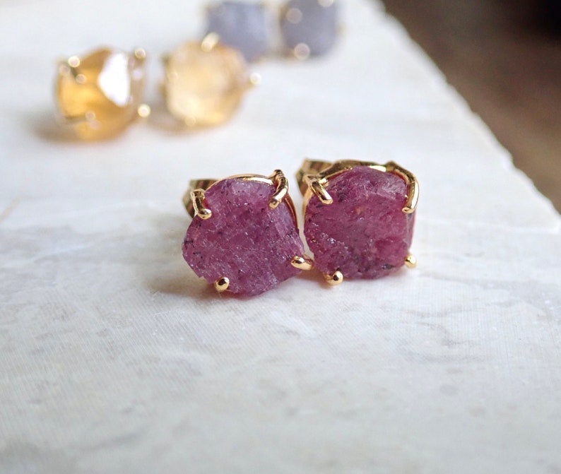 May include: A pair of stud earrings featuring raw, deep pink gemstones set in gold-toned metal prongs. In the background, additional pairs of stud earrings with yellow and light blue raw gemstones are visible, each secured with gold-toned prongs.
