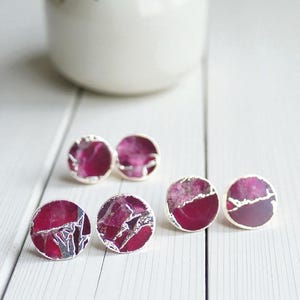 May include: Six pairs of round stud earrings with a silver border. The earrings have a deep magenta centre with a marbled, textured appearance. A white vase with a sprig of greenery is in the background.