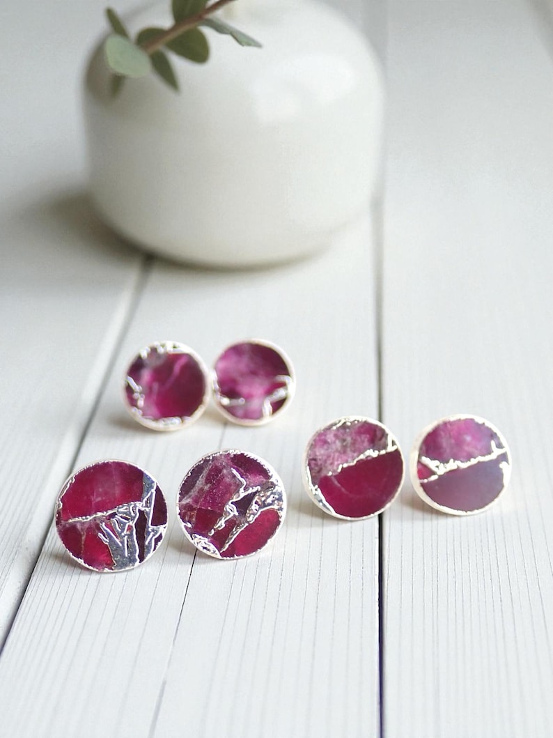 May include: Six pairs of round stud earrings with a silver border. The earrings have a deep magenta centre with a marbled, textured appearance. A white vase with a sprig of greenery is in the background.