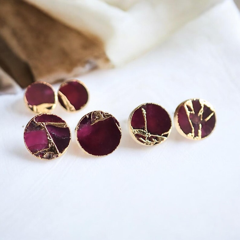 May include: Six round stud earrings with a deep burgundy centre and gold-coloured, irregular edges. The earrings are displayed on a white surface, with a soft, blurred background. The earrings are a jewellery item.