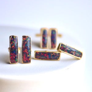 May include: A collection of gold-toned jewelry featuring rectangular black opal stones with red, green, and blue flecks. The set includes a ring and several pairs of earrings.