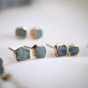 May include: A set of four gold-rimmed earrings featuring raw, irregular-shaped blue-green gemstones. The earrings are a minimalist style and would make a great gift.