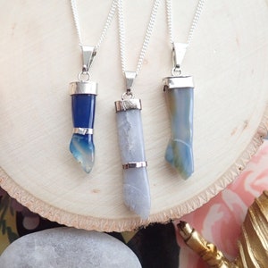 May include: Three silver necklaces with blue agate pendants. The pendants are all different shapes, with one being a long, thin rectangle, one being a short, wide rectangle, and one being a curved shape. The necklaces are all on a white wooden surface.