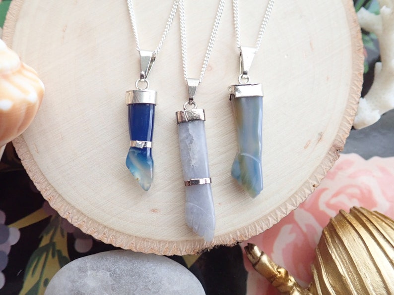 May include: Three silver necklaces with blue agate pendants. The pendants are all different shapes, with one being a long, thin rectangle, one being a short, wide rectangle, and one being a curved shape. The necklaces are all on a white wooden surface.