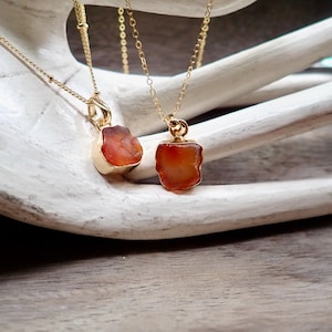 May include: Two gold necklaces with carnelian stone pendants. The carnelian stones are a deep orange color and have a rough, natural texture. The necklaces are delicate and would make a beautiful gift.