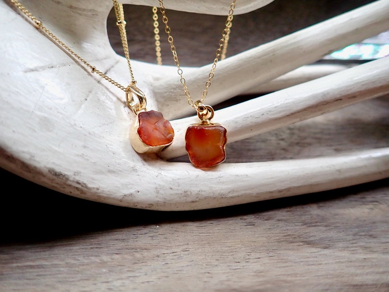 May include: Two gold necklaces with carnelian stone pendants. The carnelian stones are a deep orange color and have a rough, natural texture. The necklaces are delicate and would make a beautiful gift.