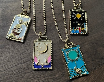 May include: Four gold-colored pendant necklaces with tarot card designs. Each rectangular pendant features a different colorful illustration and is suspended from a gold chain. The designs include a skeleton, a crescent moon, a sun, and a starry night.