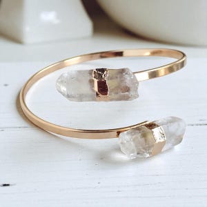 May include: A gold-colored, open cuff bracelet featuring two clear quartz crystal stones. The crystals are secured with gold-toned metal accents. The bracelet is displayed on a white surface.