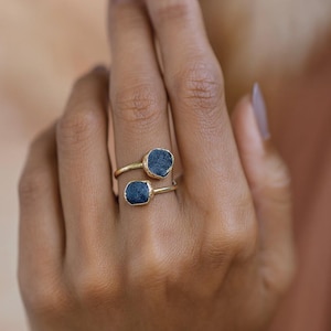 May include: A gold-toned ring featuring two dark blue, irregularly shaped gemstones. The ring's design wraps around the finger, with one stone positioned above the other. The gemstones have a textured appearance, set against the smooth, polished metal.