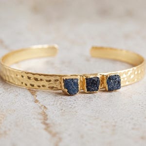 May include: A gold-toned cuff bracelet featuring a hammered texture along the band and three small, dark blue, rough-cut stones set in a row on the front. The bracelet has an open back design, allowing for adjustment to fit the wrist securely.