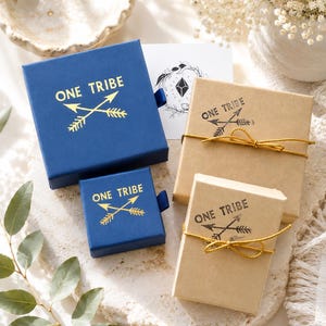 May include: Two square blue sliding jewellery boxes and two rectangular tan cardboard boxes with gold string. Each box features the text ONE TRIBE with a crossed arrow graphic. The blue boxes have gold foil printing, while the tan boxes have black ink.