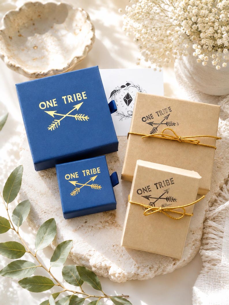 May include: Two square blue sliding jewellery boxes and two rectangular tan cardboard boxes with gold string. Each box features the text ONE TRIBE with a crossed arrow graphic. The blue boxes have gold foil printing, while the tan boxes have black ink.