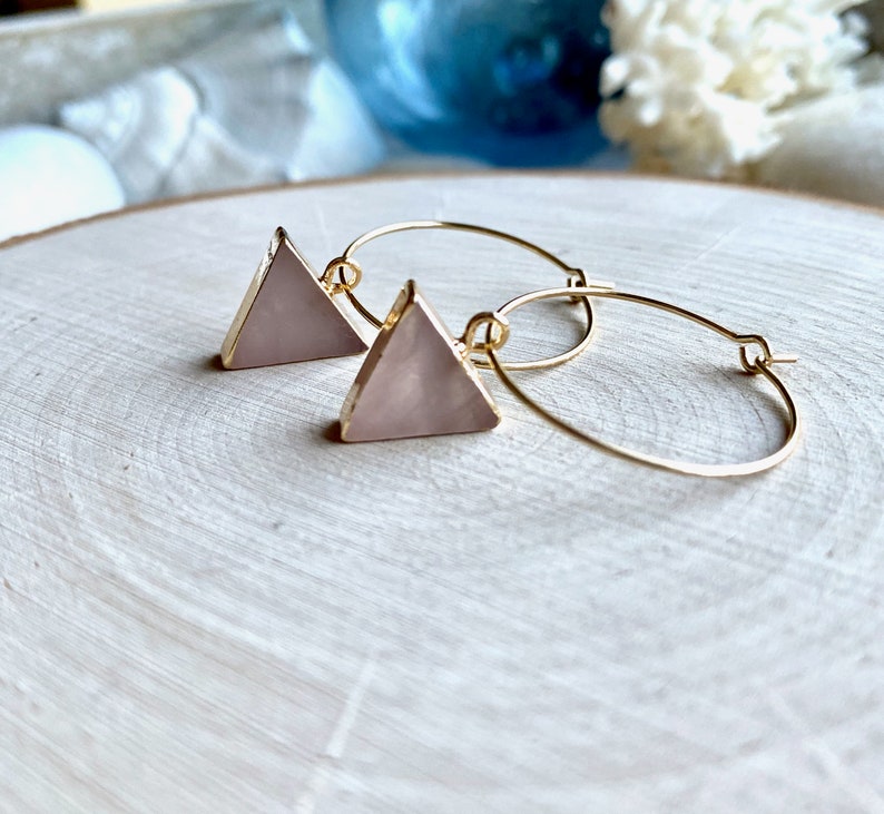 May include: Gold hoop earrings with a pink triangle pendant. The triangle is made of a translucent material with a slight shimmer.