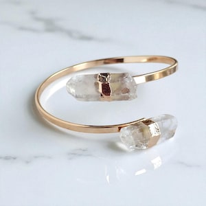 May include: A gold-coloured cuff bracelet with two clear quartz crystal accents. The crystals are secured with gold-coloured metal bands. The bracelet is displayed on a white marble surface.