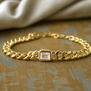 May include: A gold-colored bracelet with a curb chain design. The bracelet features a square, clear gemstone in the center. The jewelry is displayed on a textured, gold-toned surface.