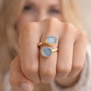 Può includere: Due anelli d'oro con pietre azzurre dalla forma irregolare. Gli anelli sono su un dito. Le pietre hanno una consistenza ruvida e naturale. Gli anelli hanno un design semplice ed elegante.