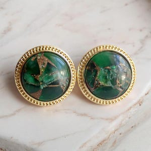 May include: A pair of round stud earrings. Each earring features a vibrant green stone with gold-colored veining, set within a gold-toned, textured frame. The earrings are displayed on a marble surface.