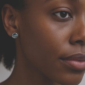 May include: Close-up of a person's face with a silver stud earring. The earring has a round shape with a blue and white design. The person has dark skin and brown eyes.