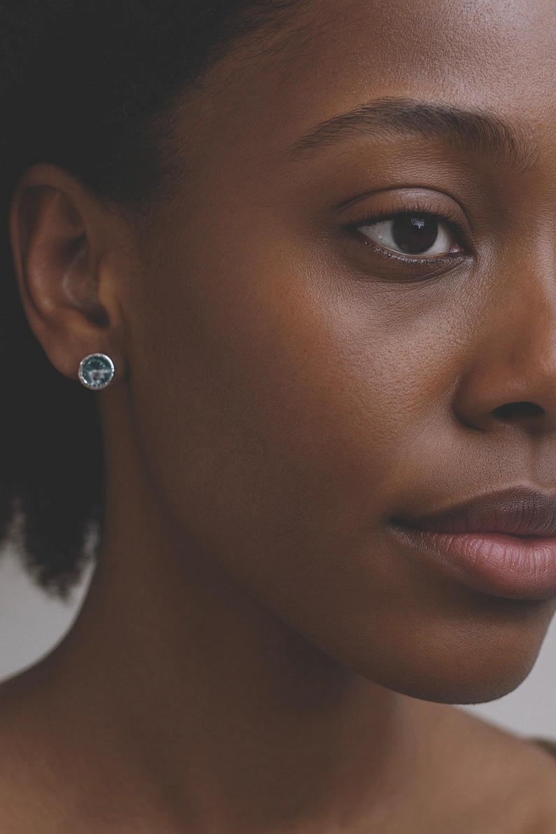 May include: Close-up of a person's face with a silver stud earring. The earring has a round shape with a blue and white design. The person has dark skin and brown eyes.