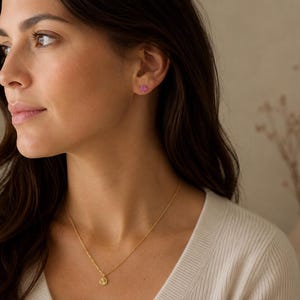 May include: A gold-toned necklace featuring a small, circular pendant with a textured surface, paired with a single small, pink, flower-shaped stud earring. The jewellery is worn against a white ribbed top, highlighting the delicate metal chain.