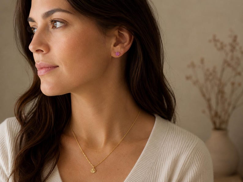 May include: A gold-toned necklace featuring a small, circular pendant with a textured surface, paired with a single small, pink, flower-shaped stud earring. The jewellery is worn against a white ribbed top, highlighting the delicate metal chain.